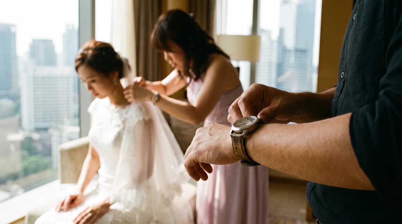 Close-up of a photographer checking the time on a wristwatch while a bride in a white gown is being pinned by a bridesmaid in a hotel suite