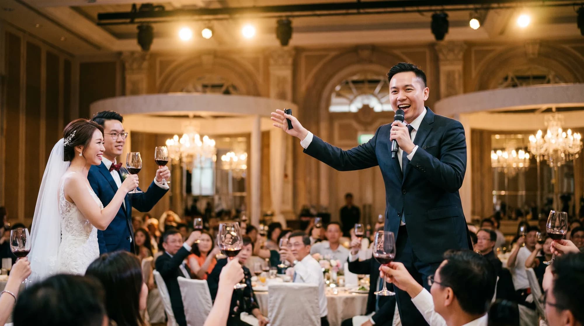 A charismatic wedding emcee in a suit holding a microphone on a ballroom stage in Singapore
