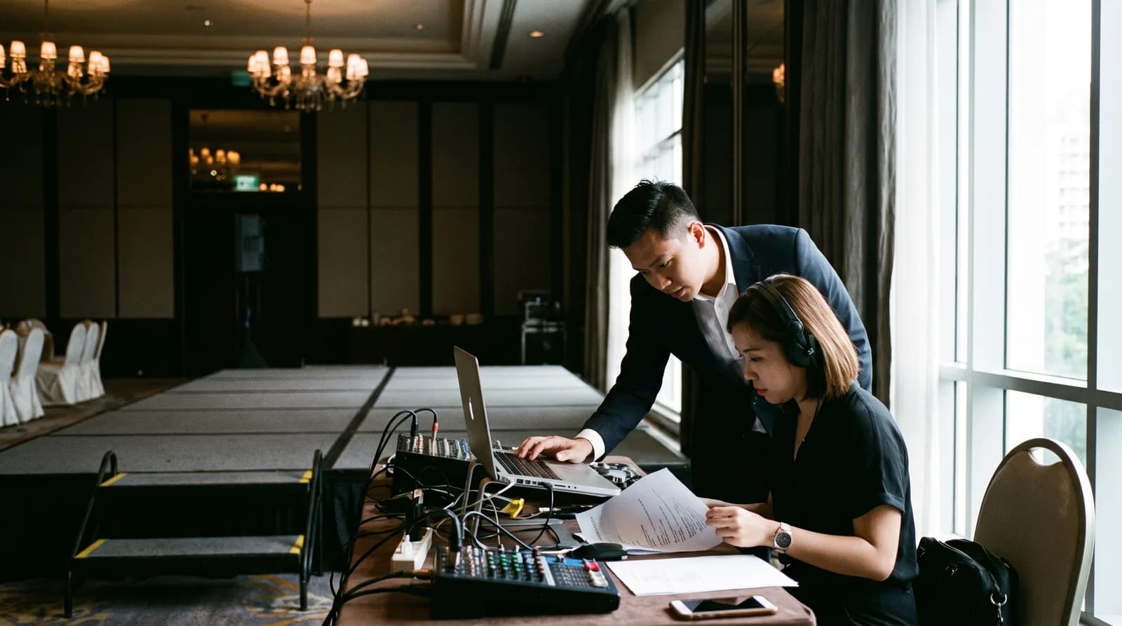 An emcee and a DJ at an AV table beside the stage checking a laptop and a running sheet before guests arrive
