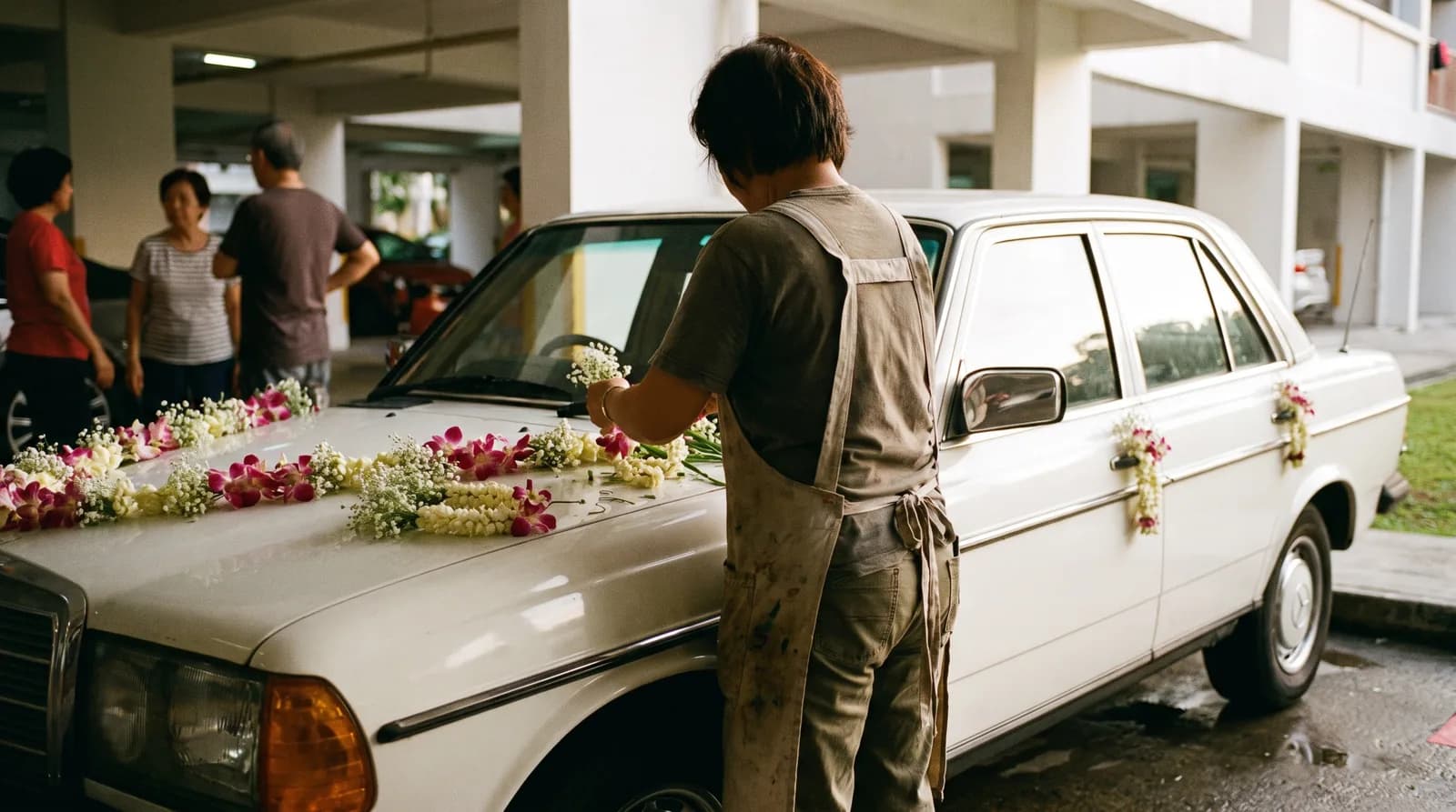 A florist fitting fresh flowers onto the bonnet and doors of a white bridal car in an HDB carpark at dusk.